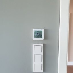 Modern System - Biały, kwadratowy termostat ścienny z wyświetlaną temperaturą 23.6 stopni Celsjusza, zamontowany nad potrójnym, białym włącznikiem światła na szarej ścianie, widoczny fragment białej framugi drzwi.