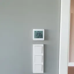 Biały, kwadratowy termostat ścienny z wyświetlaną temperaturą 23.6 stopni Celsjusza, zamontowany nad potrójnym, białym włącznikiem światła na szarej ścianie, widoczny fragment białej framugi drzwi.