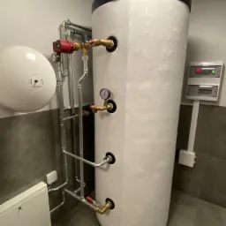 Biały, cylindryczny zasobnik ciepłej wody użytkowej z podłączonymi rurami i manometrami, zainstalowany w pomieszczeniu technicznym w Borkach, widoczna także biała skrzynka rozdzielcza i okrągły...
