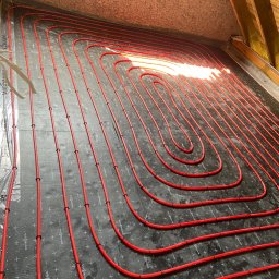 Modern System - Czerwone rury ogrzewania podłogowego, ułożone w pętle na czarnej macie izolacyjnej z widocznymi znakami producenta, instalacja w trakcie realizacji na poddaszu, widoczne elementy więźby dachowej.