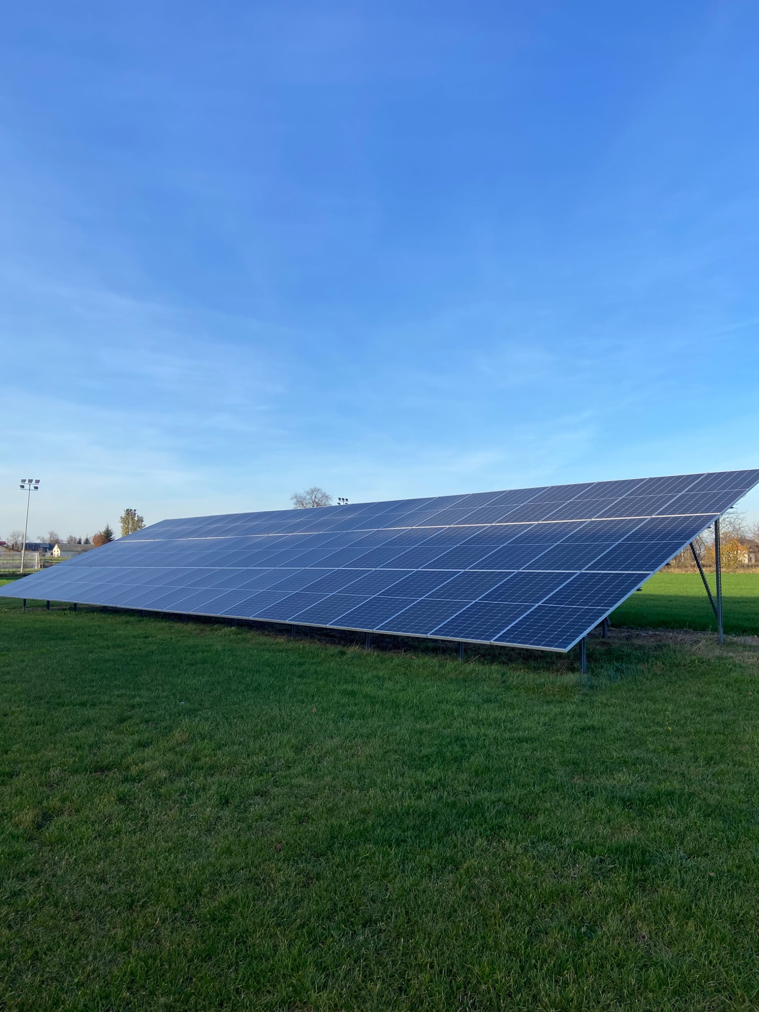 Rozległa instalacja paneli fotowoltaicznych na metalowej konstrukcji, umieszczona na trawiastym terenie, pod błękitnym niebem z delikatnymi chmurami, widoczne słupy oświetleniowe w tle.