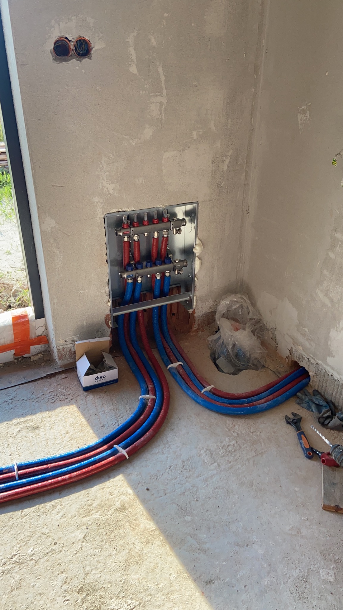 Rozdzielacz ogrzewania podłogowego z czerwono-niebieskimi rurami w surowym stanie, widoczne narzędzia i karton po akcesoriach Cluro