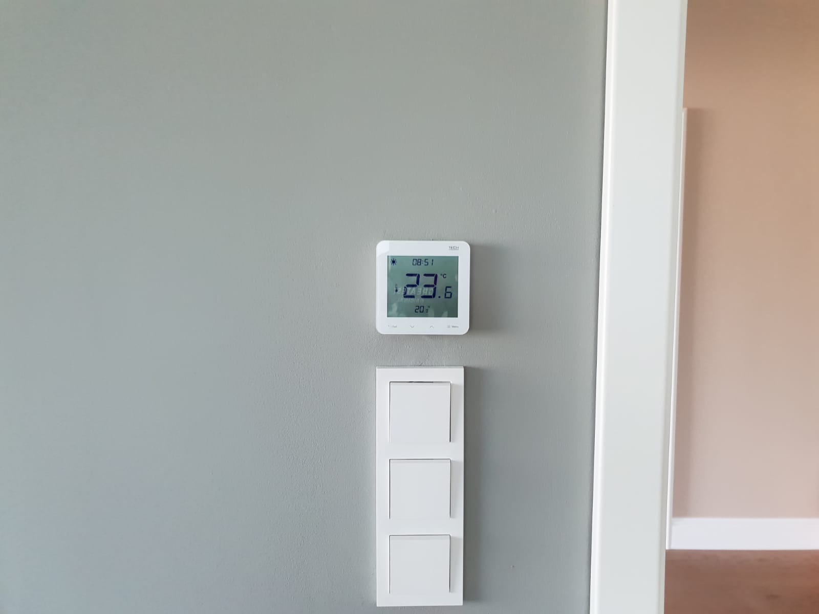 Biały, kwadratowy termostat ścienny z wyświetlaną temperaturą 23.6 stopni Celsjusza, zamontowany nad potrójnym, białym włącznikiem światła na szarej ścianie, widoczny fragment białej framugi drzwi.