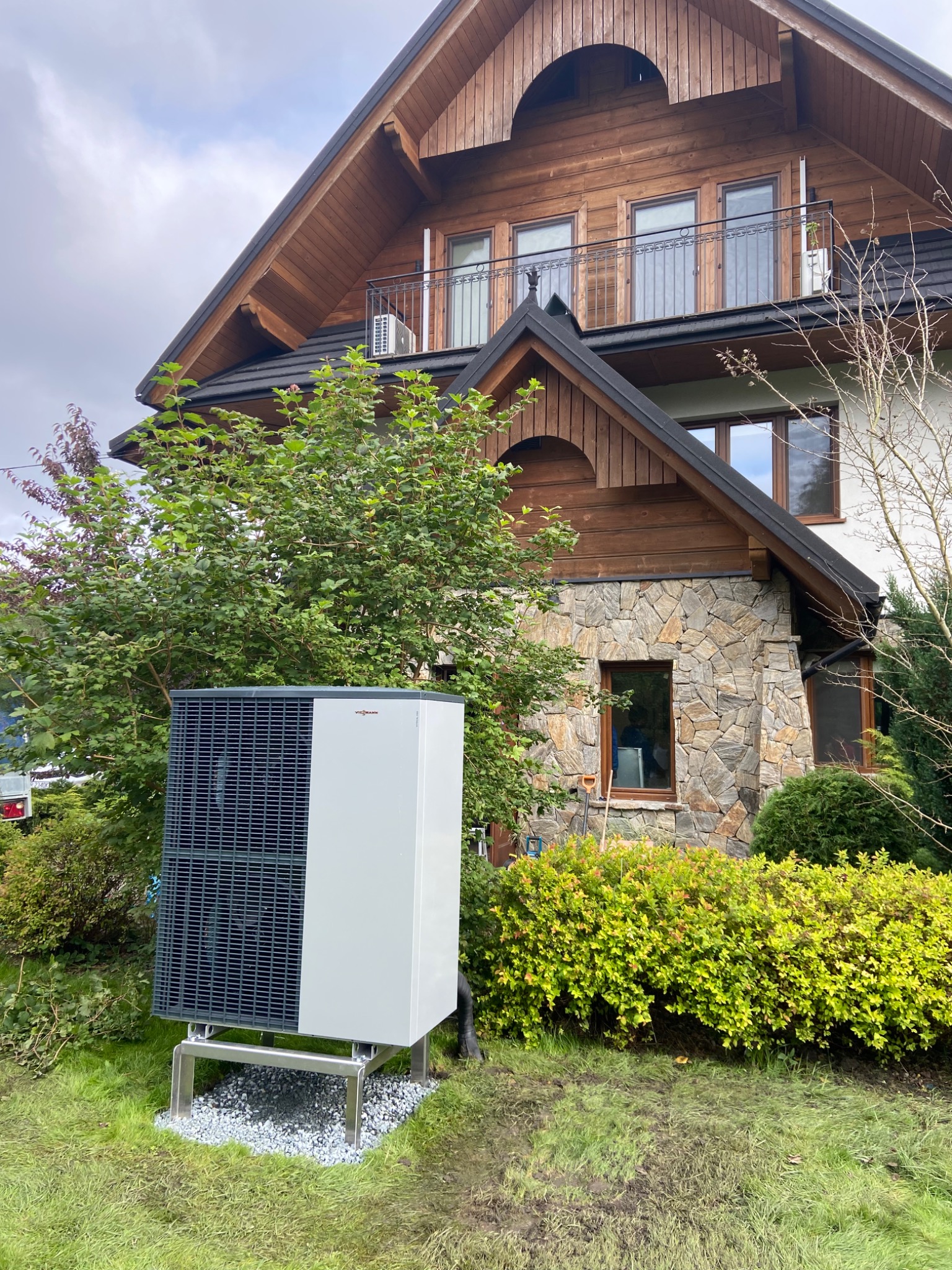 Zewnętrzna jednostka pompy ciepła Viessmann zamontowana na metalowej konstrukcji na tle domu z drewnianą elewacją i kamiennym wykończeniem, otoczona zielenią.