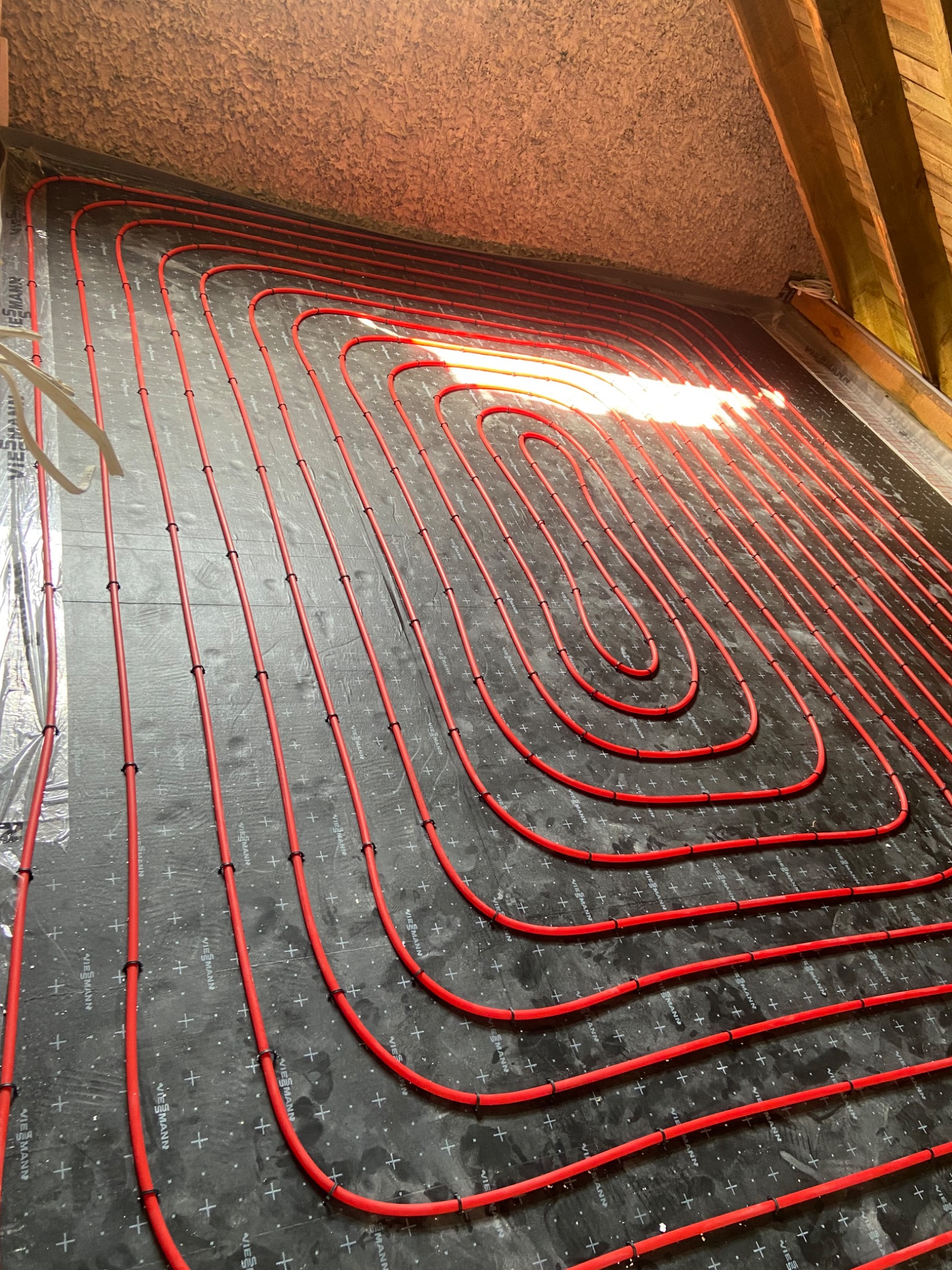 Czerwone rury ogrzewania podłogowego, ułożone w pętle na czarnej macie izolacyjnej z widocznymi znakami producenta, instalacja w trakcie realizacji na poddaszu, widoczne elementy więźby dachowej.