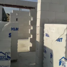 Wznoszenie ścian budynku z betonu komórkowego Solbet, widoczne palety z zapakowanymi bloczkami, w tle konstrukcja dachu sąsiedniego domu.