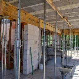 Wnętrze budynku w trakcie budowy z widocznymi ścianami z betonu komórkowego, drewnianymi belkami, stropem z prefabrykatów betonowych i metalowymi stemplami podtrzymującymi konstrukcję, w tle...