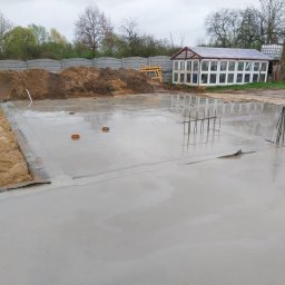 Wojtek - Wylany fundament z wystającymi prętami zbrojeniowymi, widoczny piasek i ogrodzenie betonowe w tle, obok szklarnia.