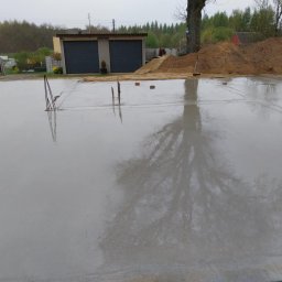 Wojtek - Świeżo wylana, mokra płyta fundamentowa z wystającymi prętami zbrojeniowymi, odbijająca drzewa i pochmurne niebo, na tle budynku gospodarczego i sterty piasku.