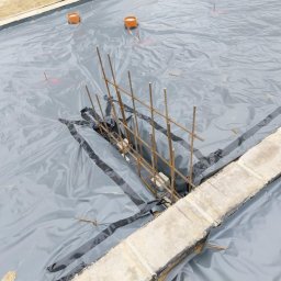 Wojtek - Przygotowanie podłoża pod fundament: szara folia izolacyjna rozłożona na piasku z wystającymi prętami zbrojeniowymi i pomarańczowymi rurami wentylacyjnymi.