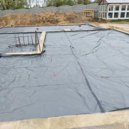 Wojtek - Przygotowany teren pod budowę: wykopany fundament, zabezpieczony szarą folią hydroizolacyjną, z widocznymi prętami zbrojeniowymi i cegłami poziomującymi, w tle hałda piasku i betonowy płot.