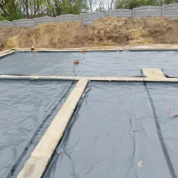 Przygotowany teren pod fundamenty z rozłożoną folią izolacyjną, widoczne betonowe ławy fundamentowe i wyjścia kanalizacyjne, w tle usypana ziemia i betonowe ogrodzenie.