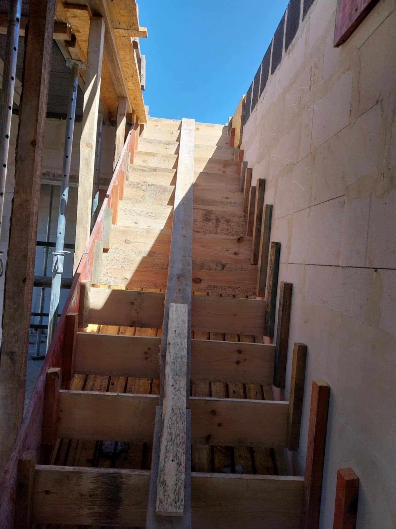 Szczegółowe ujęcie drewnianego szalunku schodów w trakcie budowy domu z bloczków, widoczne podpory i fragment ściany, niebo w tle.