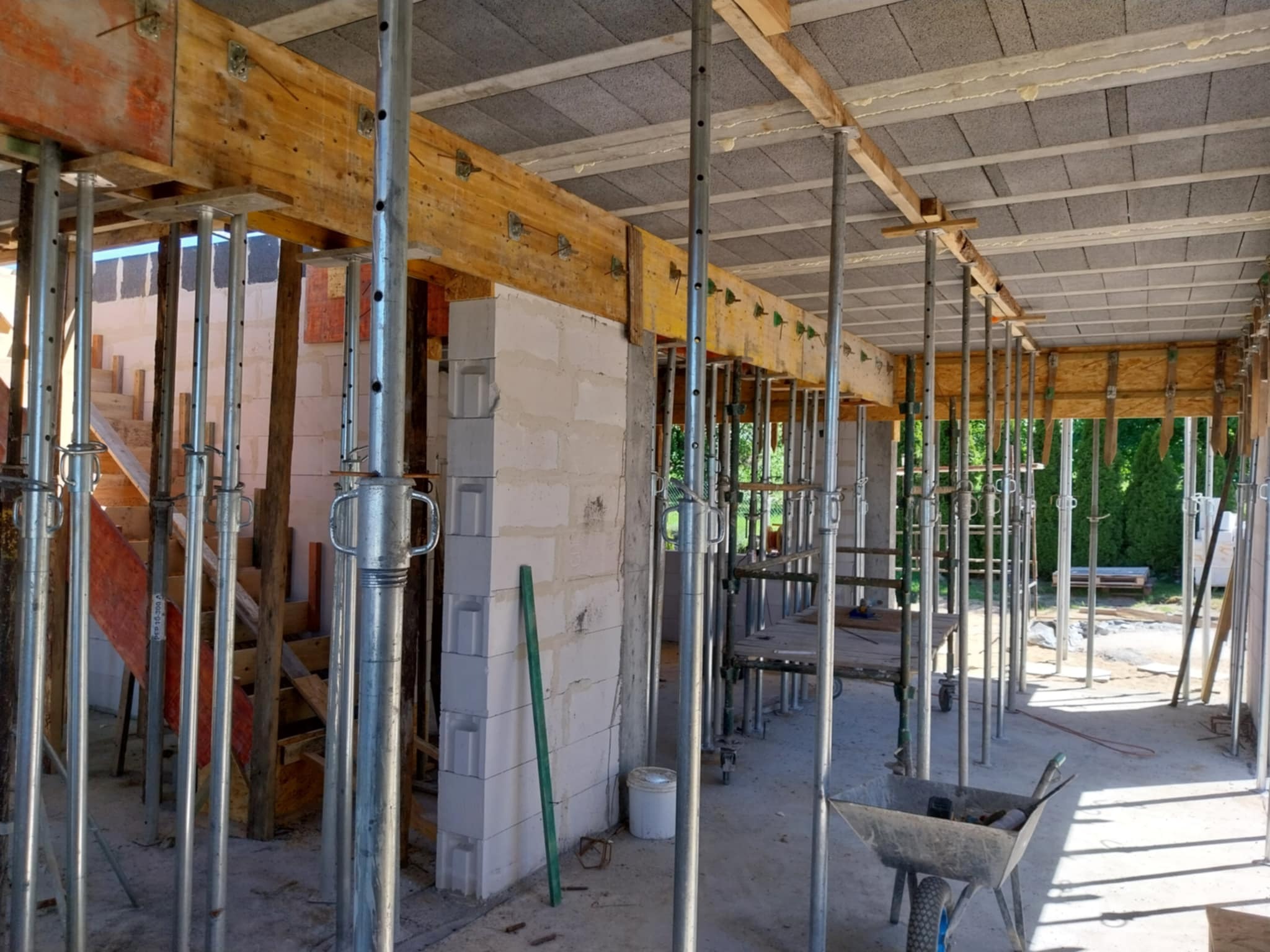 Wnętrze budynku w trakcie budowy z widocznymi ścianami z betonu komórkowego, drewnianymi belkami, stropem z prefabrykatów betonowych i metalowymi stemplami podtrzymującymi konstrukcję, w tle...