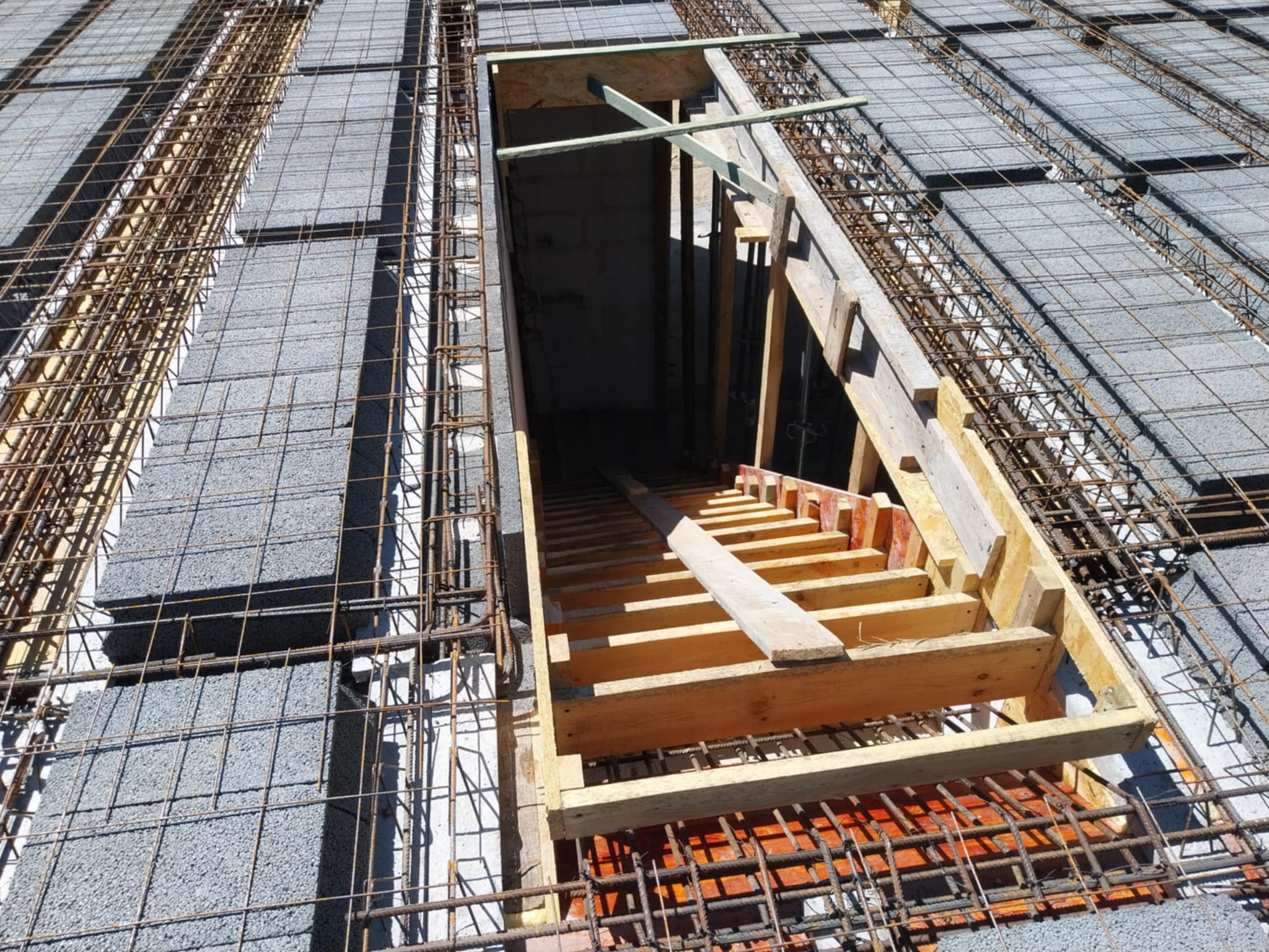 Zbrojenie stropu Teriva z pustakami betonowymi i otworem na schody tymczasowo zabezpieczonym drewnianą konstrukcją. Widoczne pręty zbrojeniowe i szare pustaki stropowe.