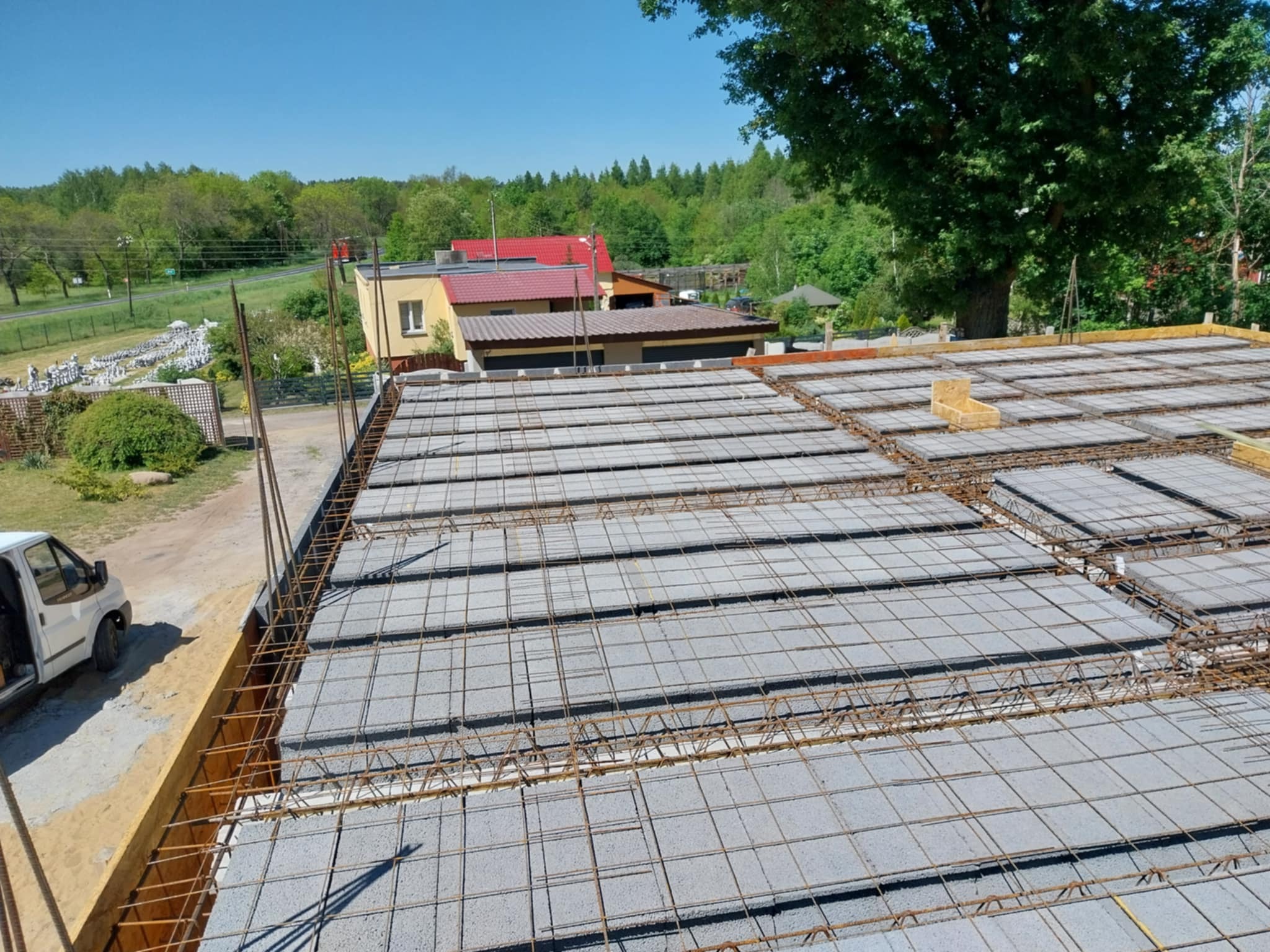 Betonowy strop Teriva z widocznym zbrojeniem stalowym przed zalaniem betonem, z deskowaniem na obrzeżach, na tle zieleni i zabudowań.