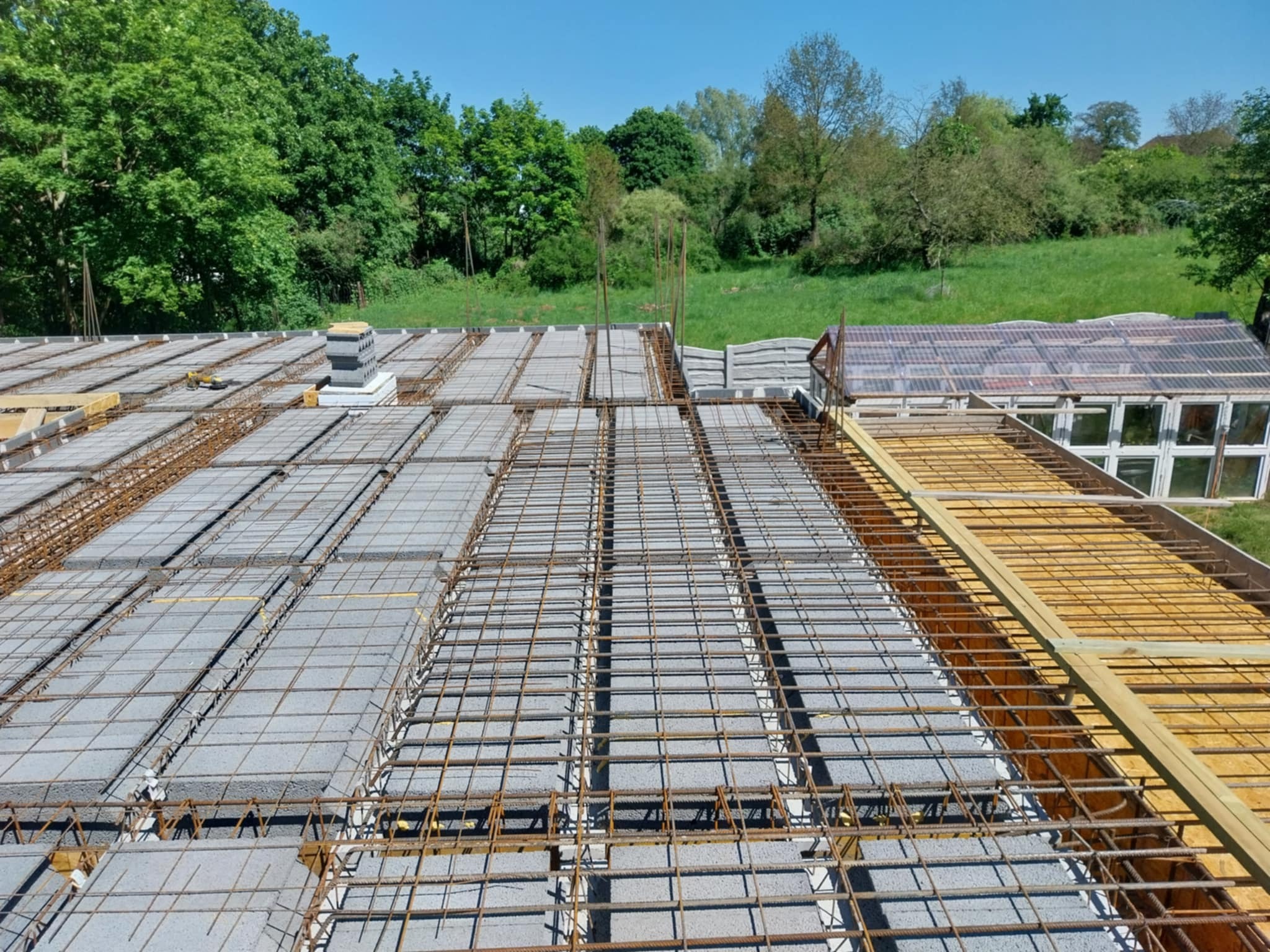 Zbrojenie stropu Teriva z pustakami betonowymi, widok z góry na siatkę zbrojeniową i drewniane szalunki, w tle zieleń drzew i budynek gospodarczy.
