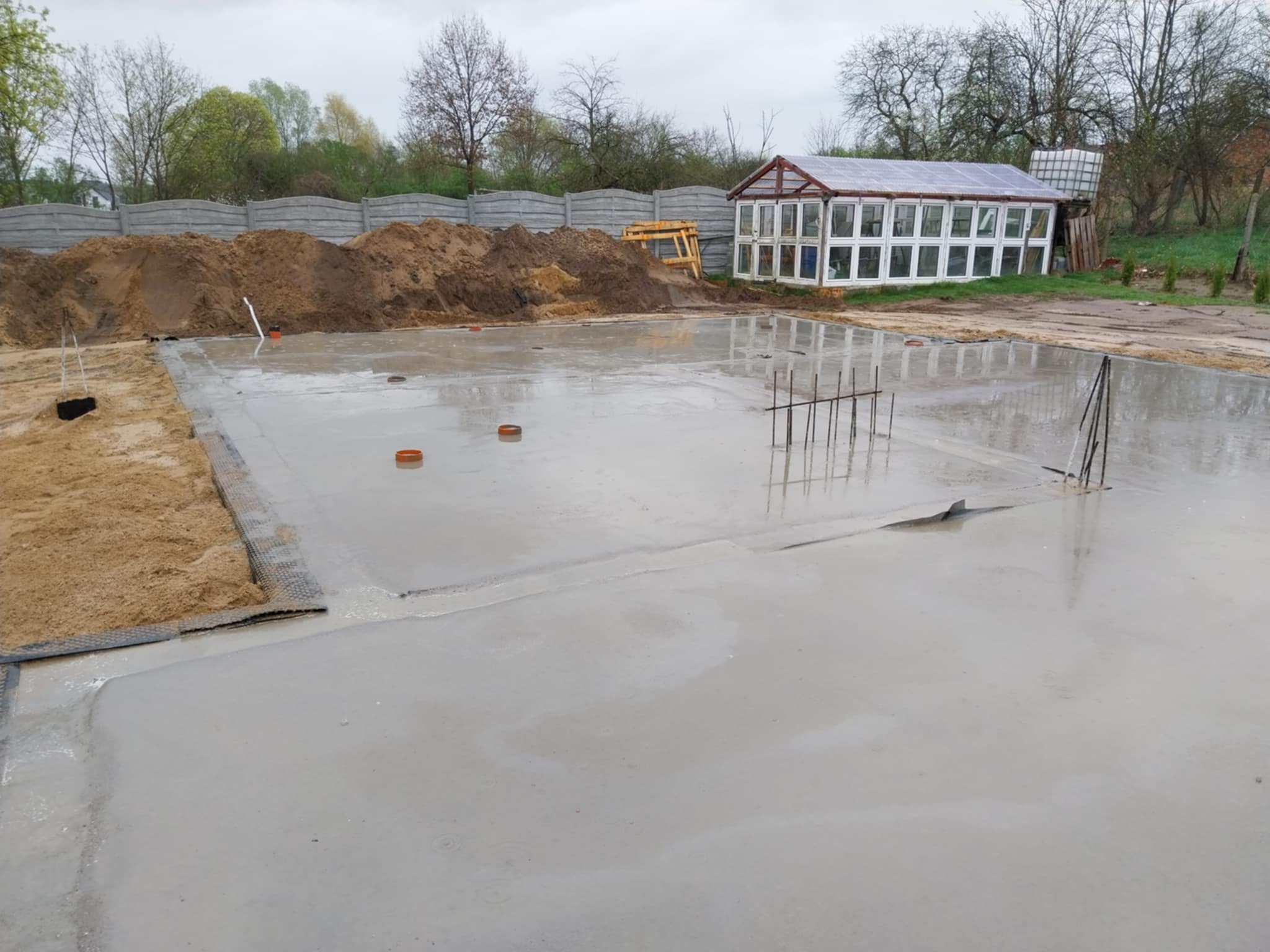 Wylany fundament z wystającymi prętami zbrojeniowymi, widoczny piasek i ogrodzenie betonowe w tle, obok szklarnia.