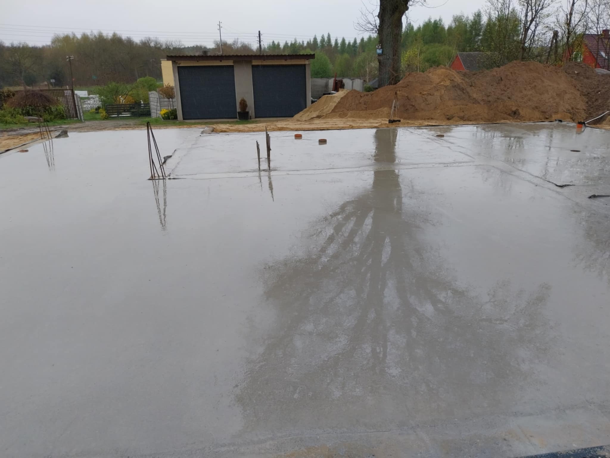 Świeżo wylana, mokra płyta fundamentowa z wystającymi prętami zbrojeniowymi, odbijająca drzewa i pochmurne niebo, na tle budynku gospodarczego i sterty piasku.