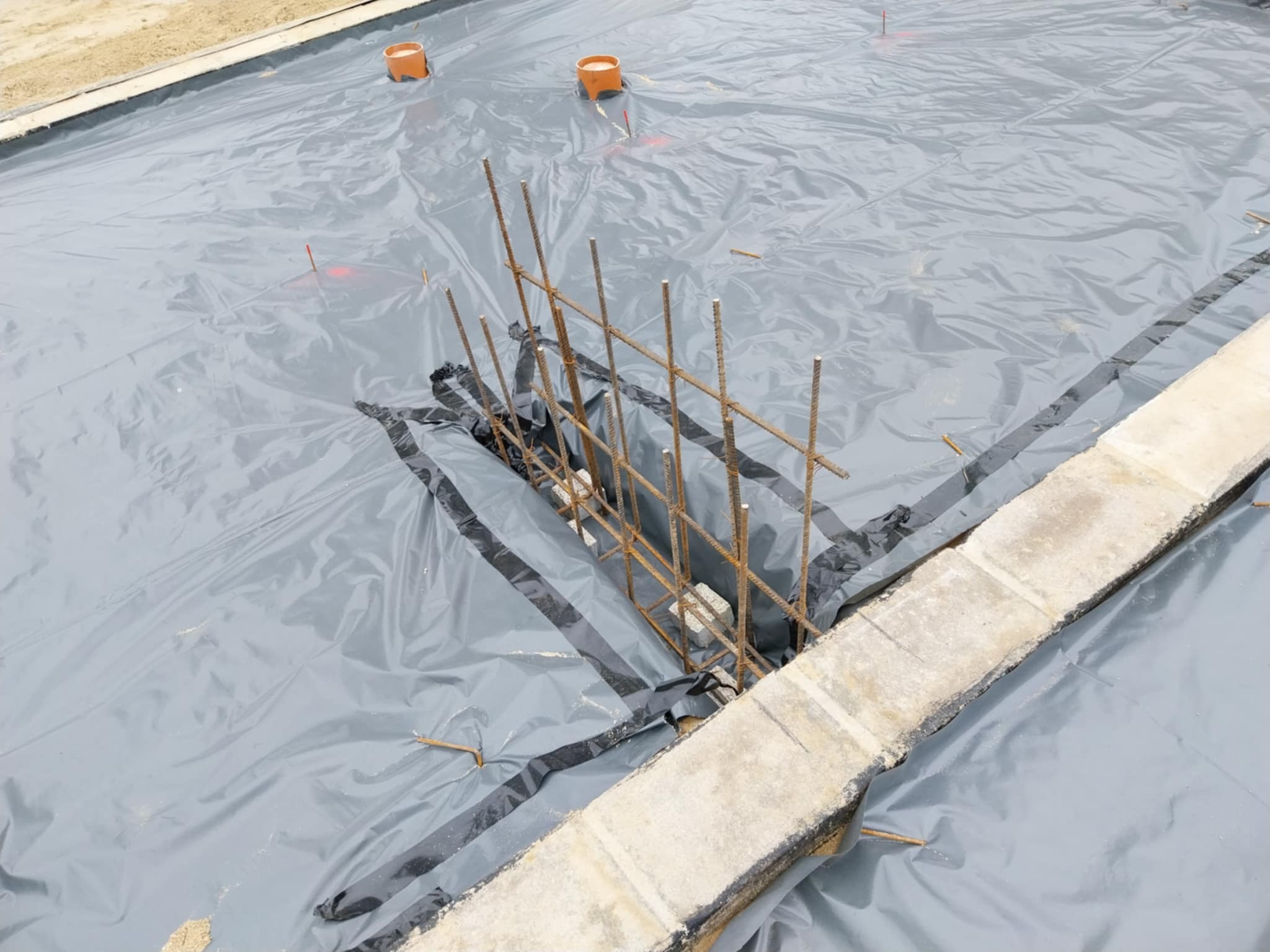 Przygotowanie podłoża pod fundament: szara folia izolacyjna rozłożona na piasku z wystającymi prętami zbrojeniowymi i pomarańczowymi rurami wentylacyjnymi.