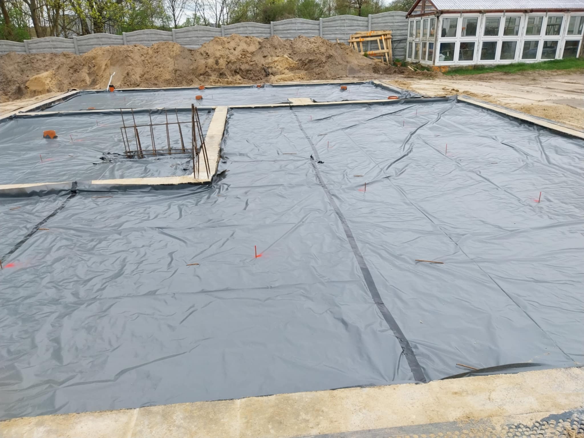 Przygotowany teren pod budowę: wykopany fundament, zabezpieczony szarą folią hydroizolacyjną, z widocznymi prętami zbrojeniowymi i cegłami poziomującymi, w tle hałda piasku i betonowy płot.