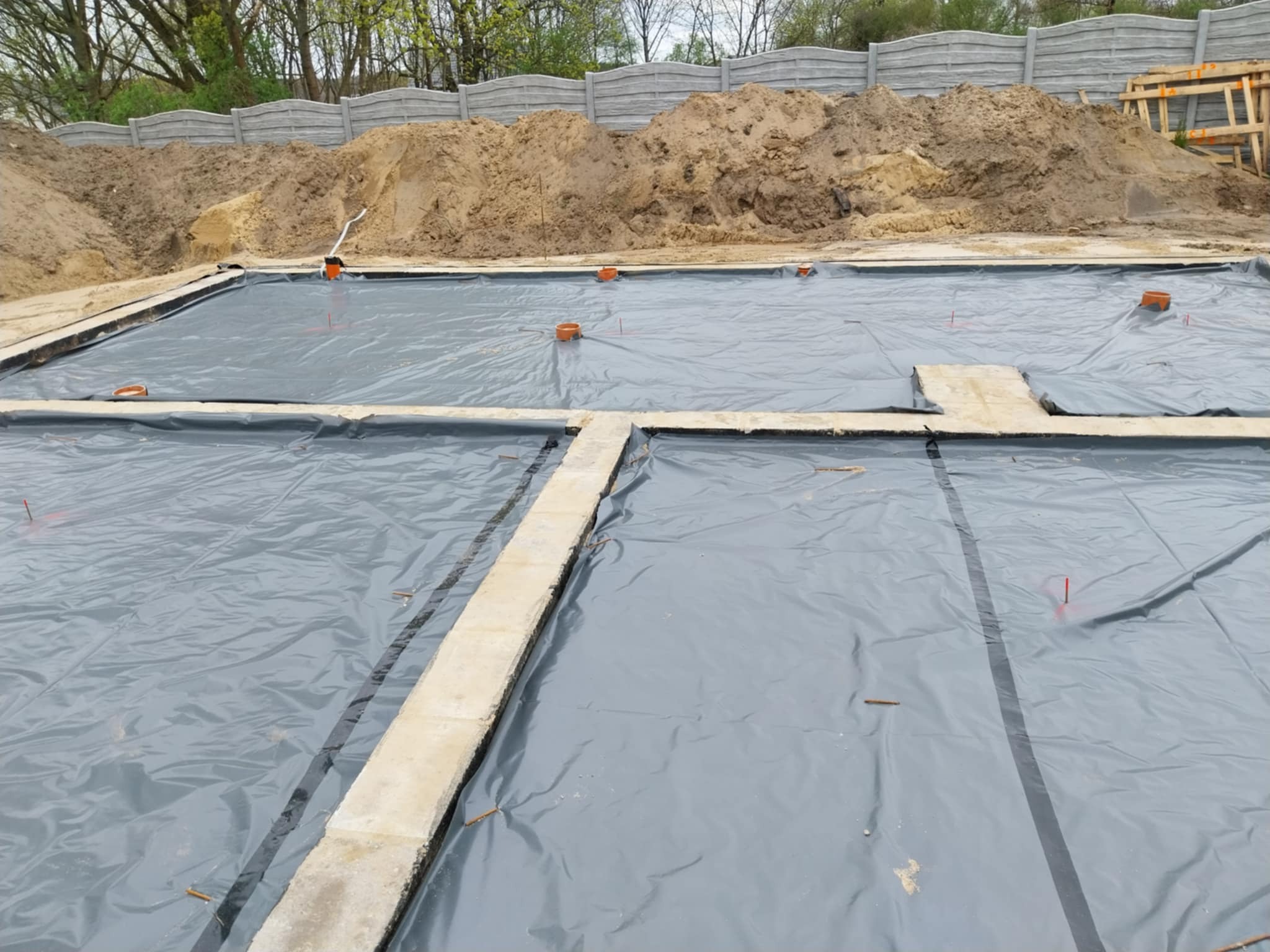 Przygotowany teren pod fundamenty z rozłożoną folią izolacyjną, widoczne betonowe ławy fundamentowe i wyjścia kanalizacyjne, w tle usypana ziemia i betonowe ogrodzenie.