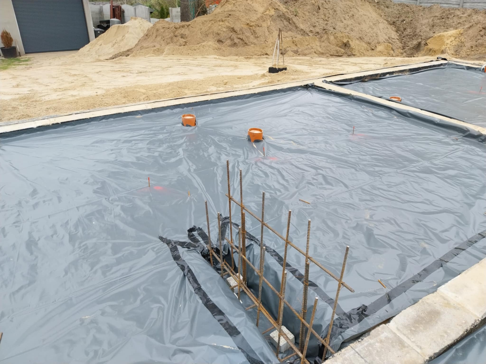 Przygotowanie terenu pod fundamenty: folia izolacyjna rozłożona na gruncie, widoczne zbrojenia i rury kanalizacyjne, w tle hałda piasku.