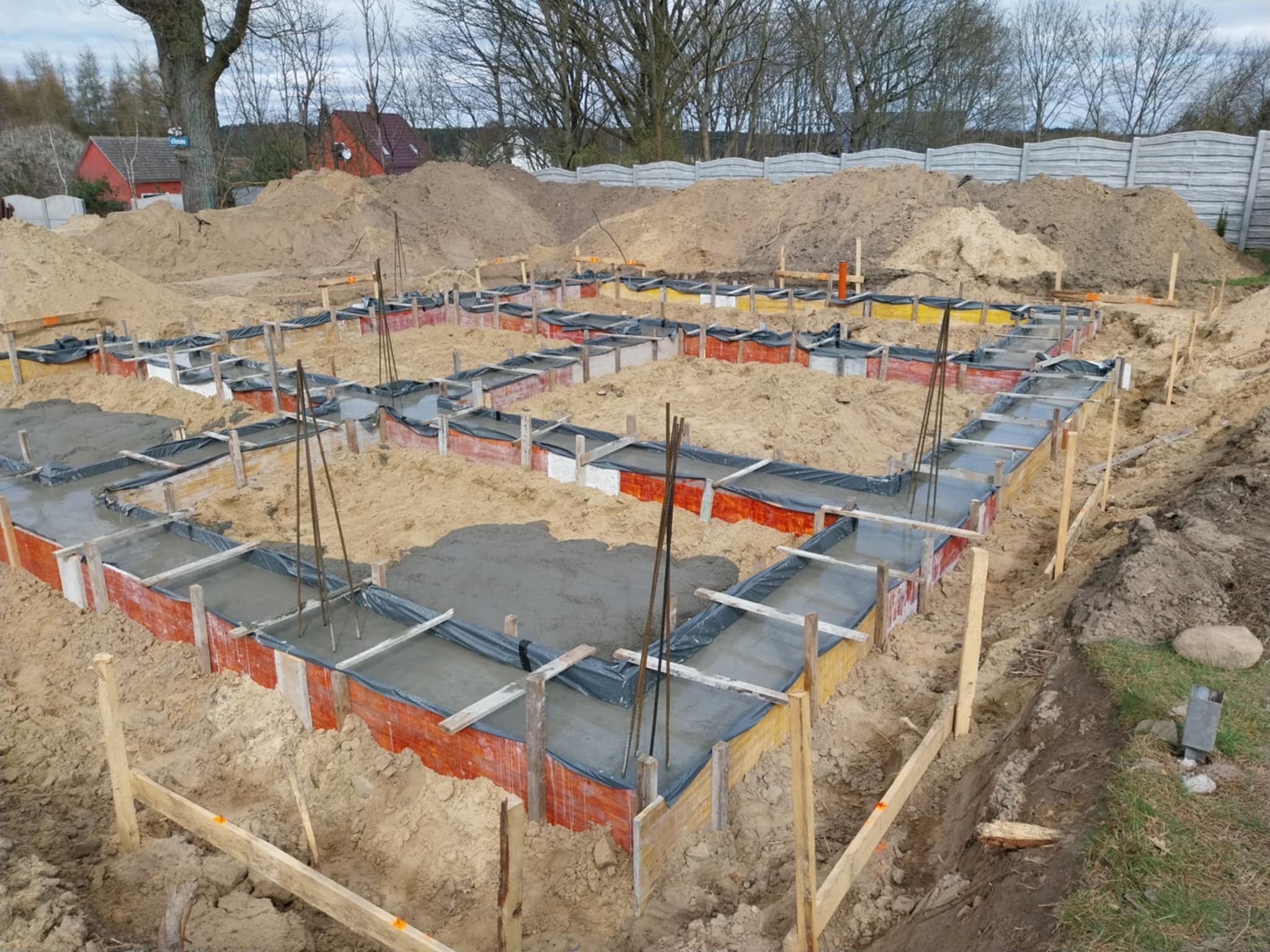 Wylany beton w szalunkach fundamentowych, zabezpieczonych folią, z wystającymi prętami zbrojeniowymi, otoczony ziemią i stertą piasku, w tle widoczny dom i drzewa.