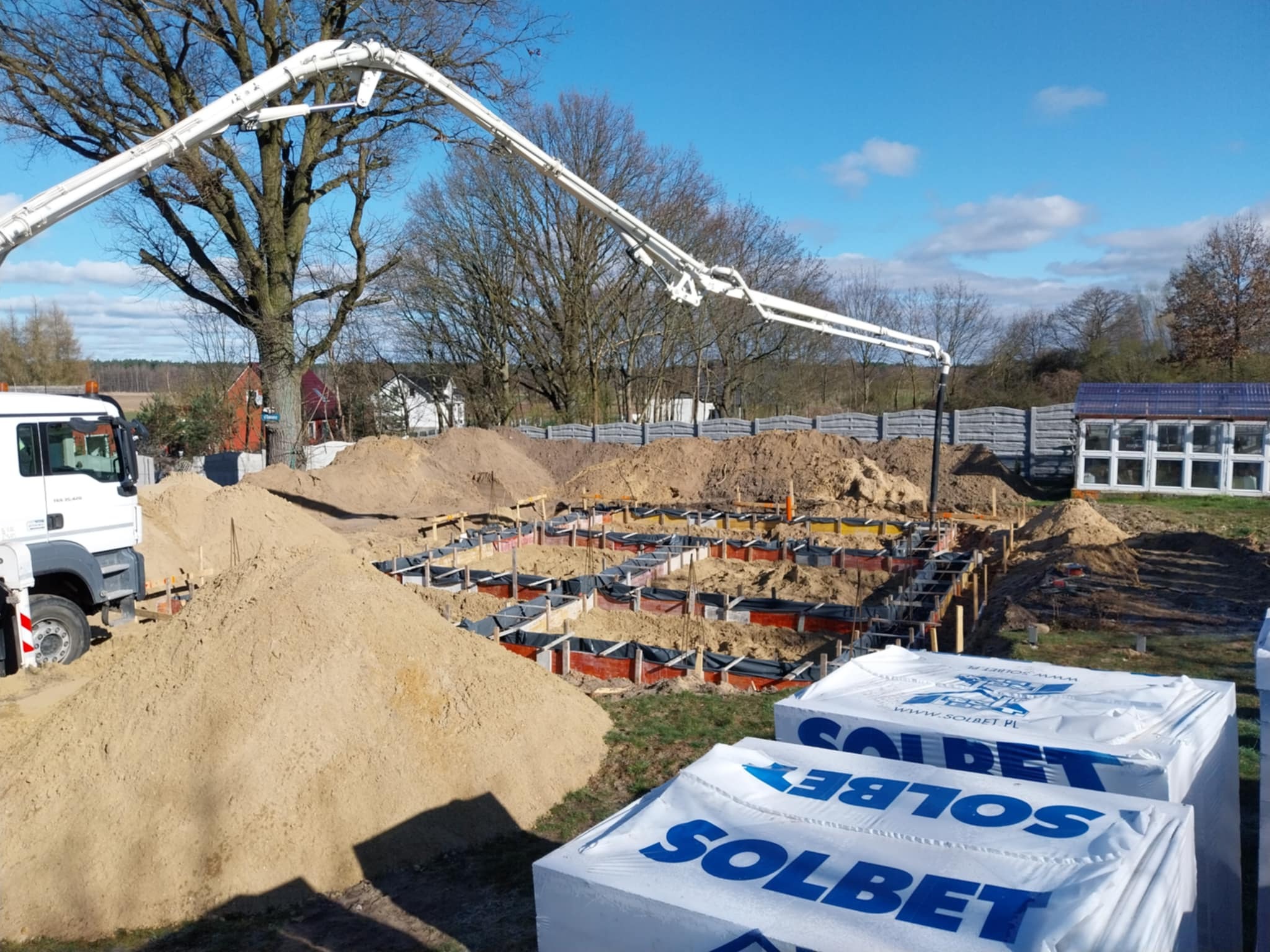 Wylewanie betonu do szalunków fundamentowych przy użyciu pompy, widoczne hałdy piasku i zapasy bloczków Solbet na tle drzew i błękitnego nieba.