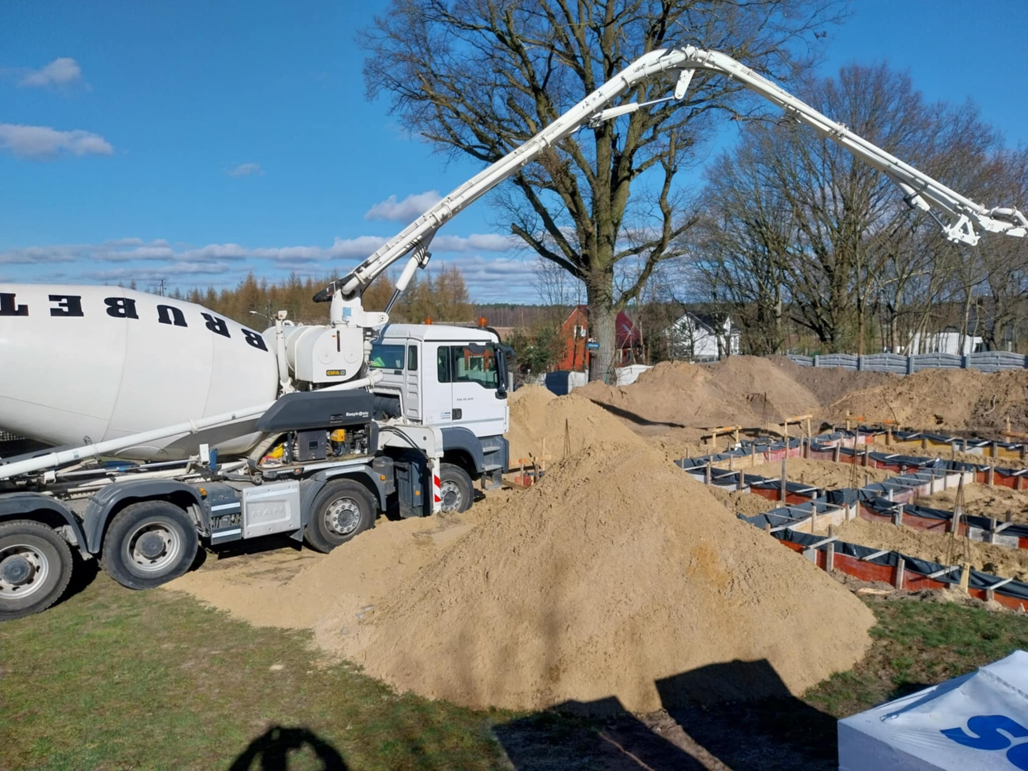 Betonomieszarka z wysięgnikiem wylewa beton do szalunków fundamentowych pod budowę domu jednorodzinnego, widoczny stos piasku.