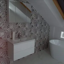 Ukośna łazienka na poddaszu z wanną wolnostojącą, umywalką z szafką i lustrem, wykończona wzorzystymi płytkami ceramicznymi w odcieniach szarości i brązu. Okno dachowe w drewnianej ramie w tle.