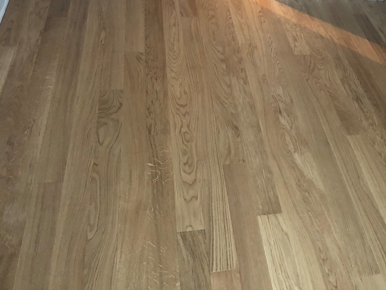 Jasna podłoga z drewnianych desek z wyraźnym usłojeniem, oświetlona z prawej strony, widoczne drobne rysy na powierzchni.