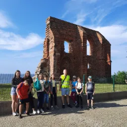 Grupa dzieci i dorosły w odblaskowej kamizelce stojący przed ruinami ceglanej budowli na tle błękitnego nieba podczas wycieczki.