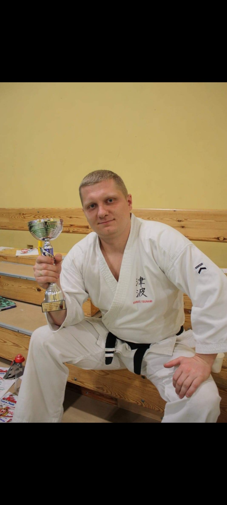 Mężczyzna w białym kimonie z czarnym pasem siedzi i trzyma srebrny puchar z plakietką, tło stanowi żółta ściana i drewniana ławka.