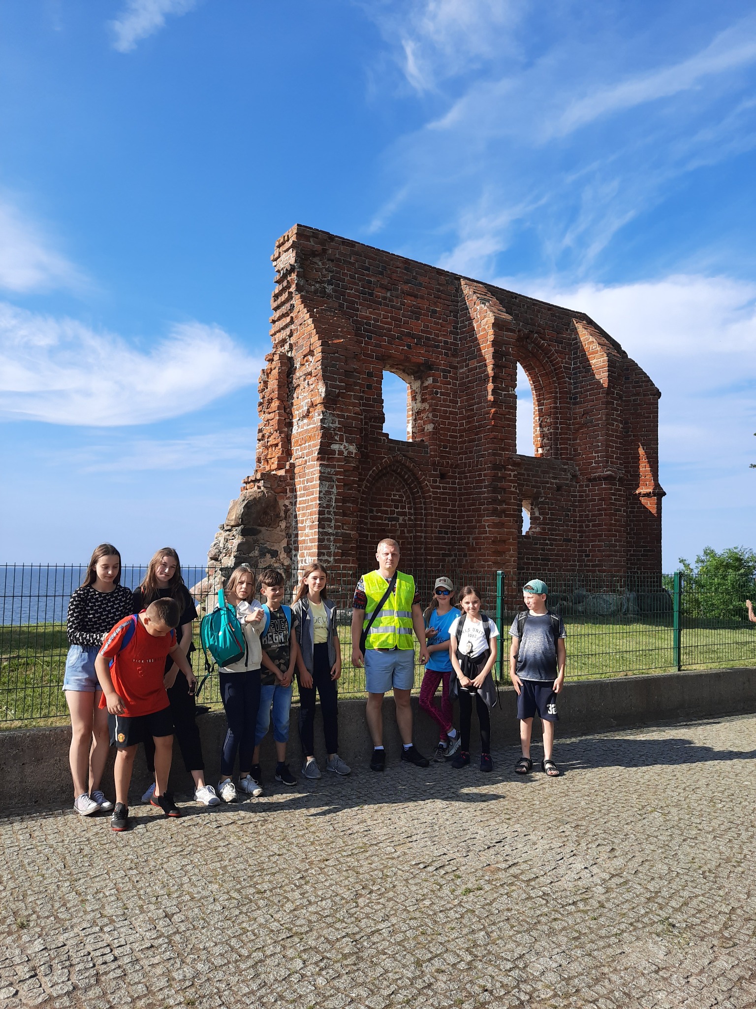 Grupa dzieci i dorosły w odblaskowej kamizelce stojący przed ruinami ceglanej budowli na tle błękitnego nieba podczas wycieczki.