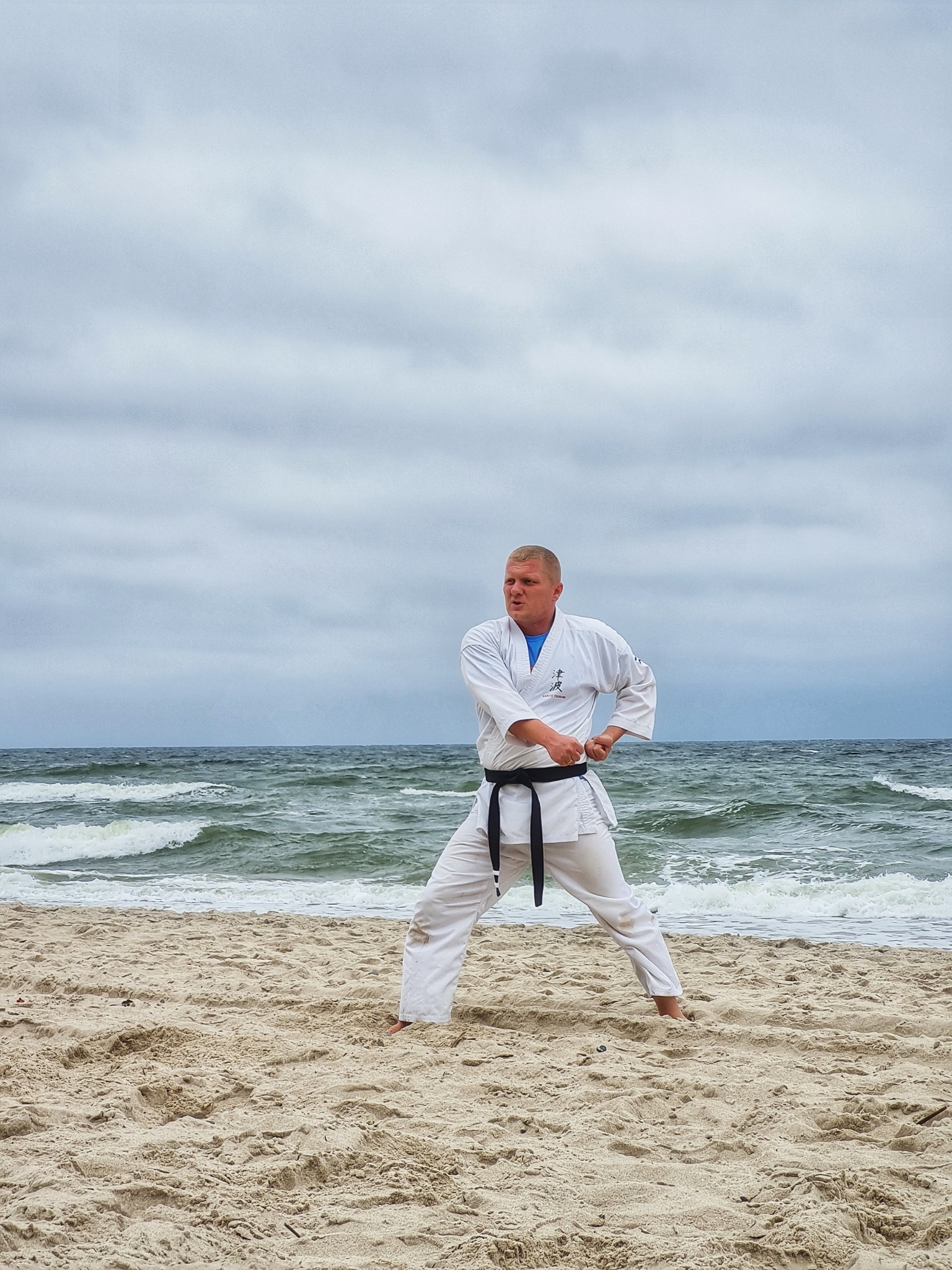 Mężczyzna w białym kimonie z czarnym pasem, trenujący karate na piaszczystej plaży nad morzem, pochmurne niebo w tle.