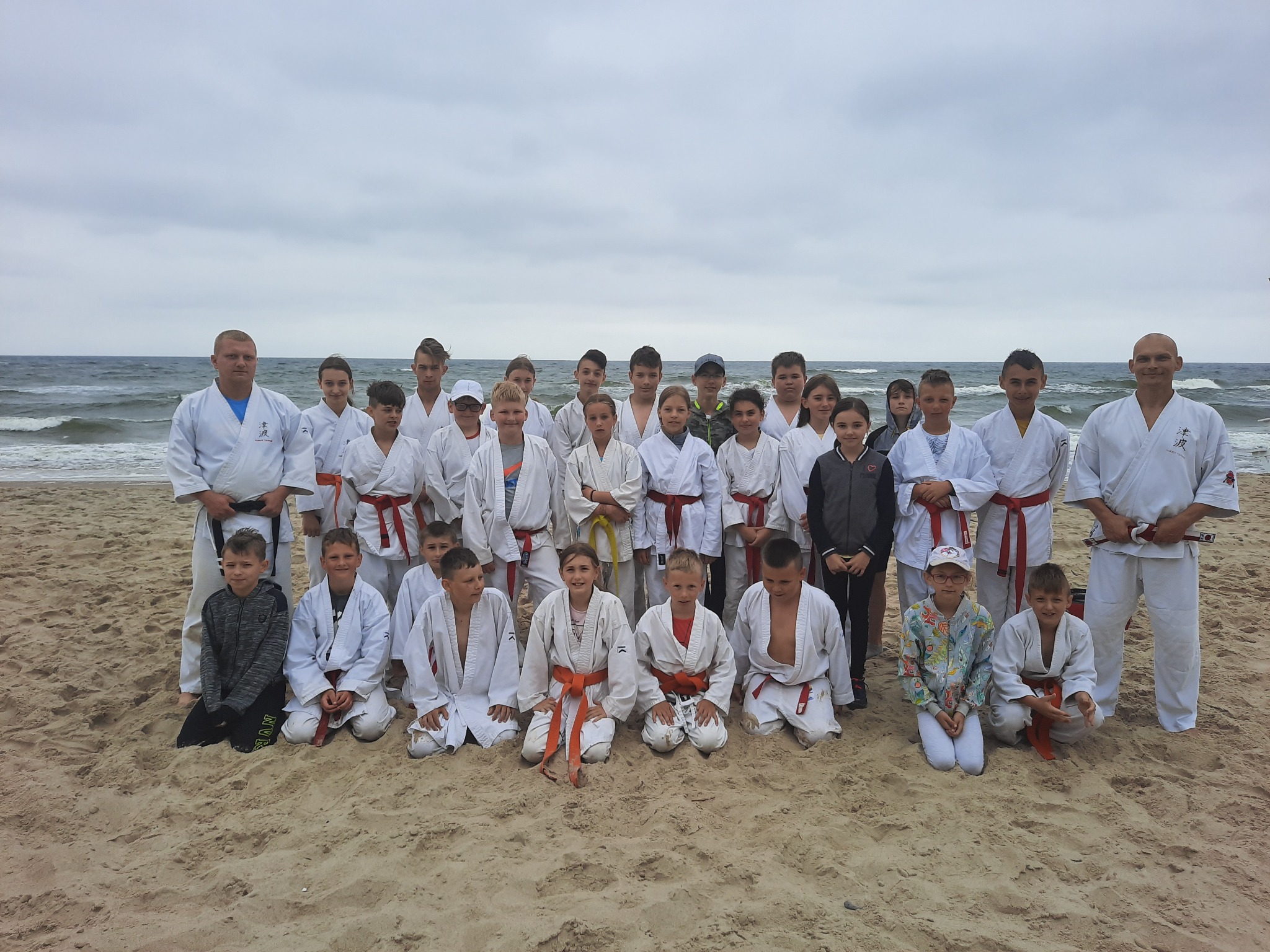 Grupa dzieci i dorosłych w białych strojach karate z różnymi kolorami pasów, ustawiona na piaszczystej plaży z widokiem na morze i pochmurne niebo.