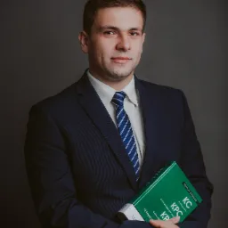 Portret mężczyzny w garniturze, trzymającego zielony podręcznik 'Kodeks Cywilny KPC' na szarym tle.