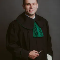 Uśmiechnięty mężczyzna w todze z zielonym żabotem trzymający długopis i kodeks na szarym tle.