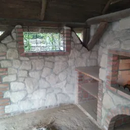 Murowana altana z kamienia i cegły, z oknem z ozdobną kratą i wnęką na grill, surowy stan podłoża.