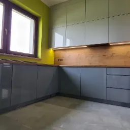 Nowoczesna zabudowa kuchenna w odcieniach szarości z drewnianym blatem i panelem ściennym, oświetlona listwą LED, na tle zielonej ściany i marmurowej podłogi.