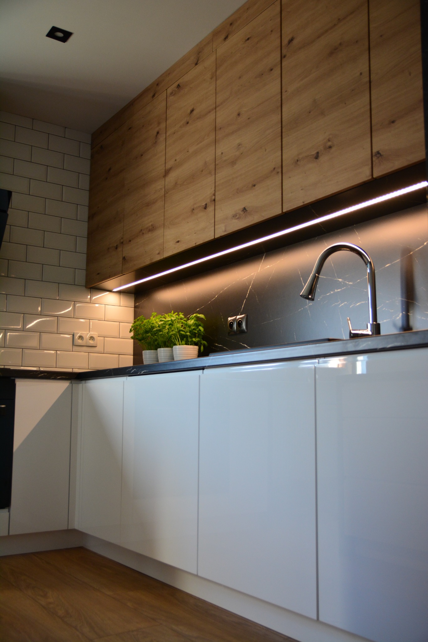 Nowoczesna kuchnia z białymi, lakierowanymi szafkami dolnymi i drewnianymi szafkami górnymi, podświetlona listwą LED, z czarnym blatem i białymi płytkami na ścianie.