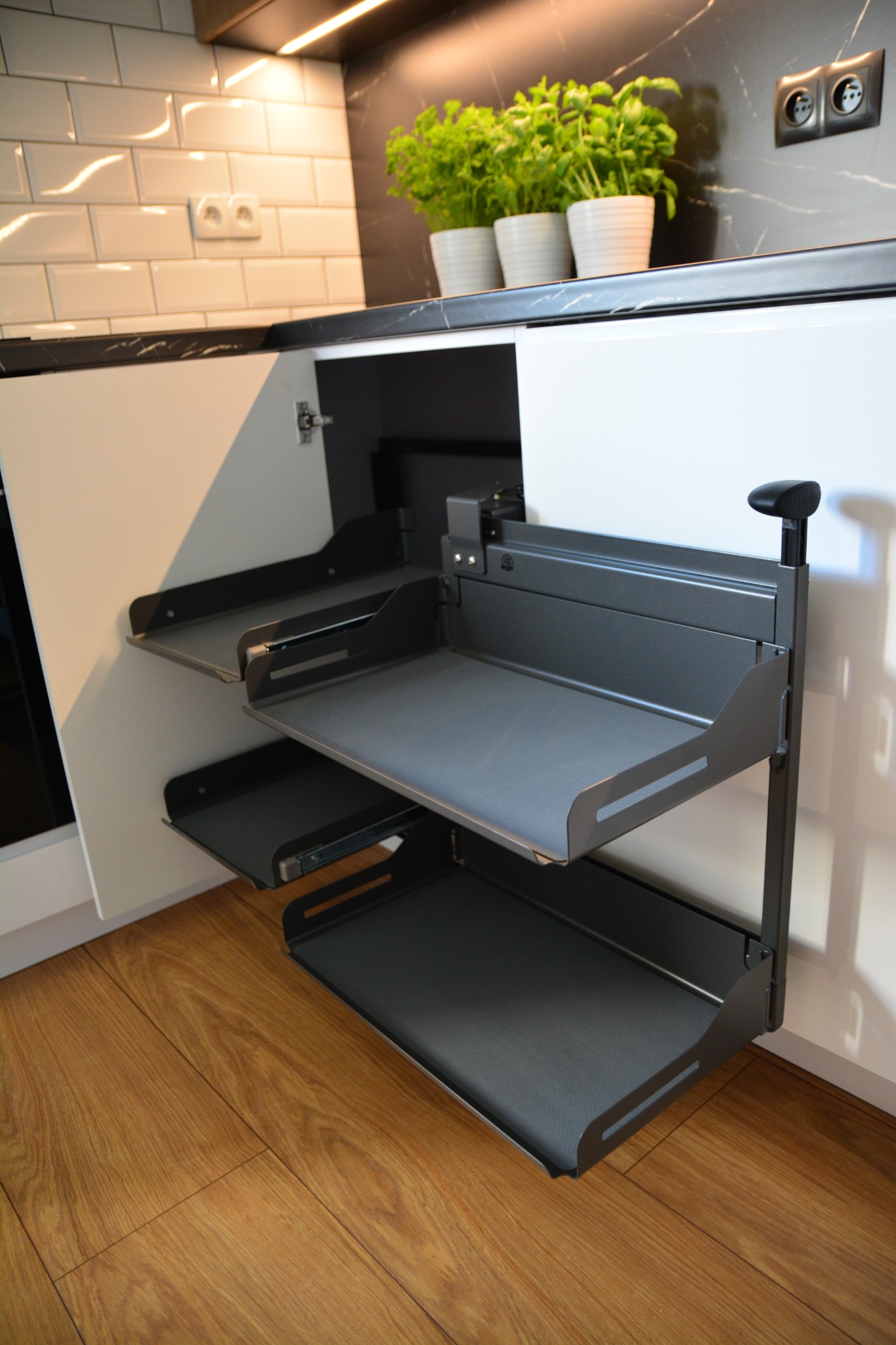 Wysuwany, trzypoziomowy system półek cargo do szafki kuchennej, z szarą, teksturowaną powierzchnią, zamontowany w białej szafce pod blatem z czarnego marmuru, widoczne doniczki z ziołami.