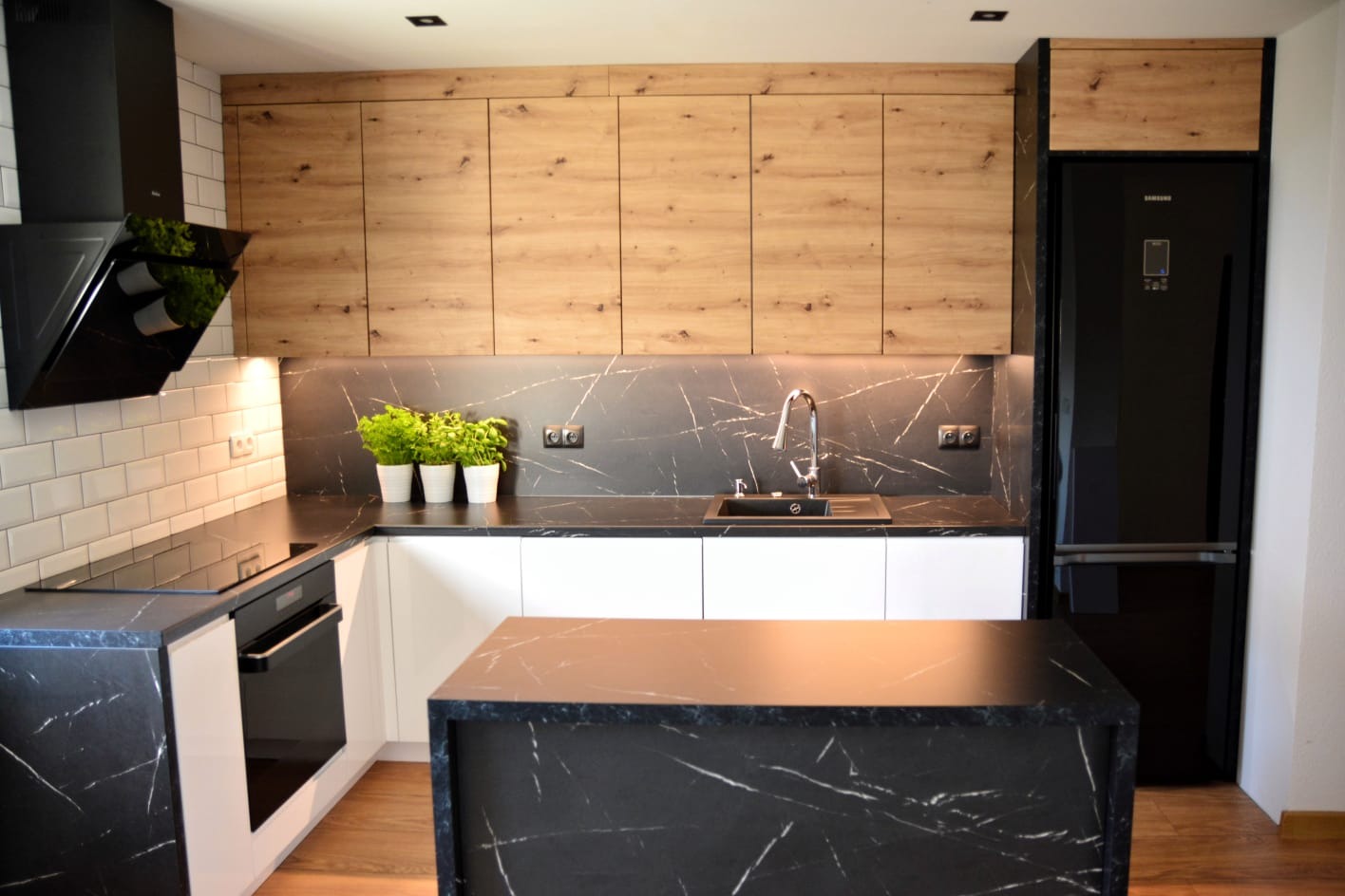 Nowoczesna kuchnia z drewnianymi szafkami górnymi, czarnym blatem imitującym marmur, wyspą kuchenną i białymi szafkami dolnymi, oświetlona punktowo, z czarnym okapem i lodówką.