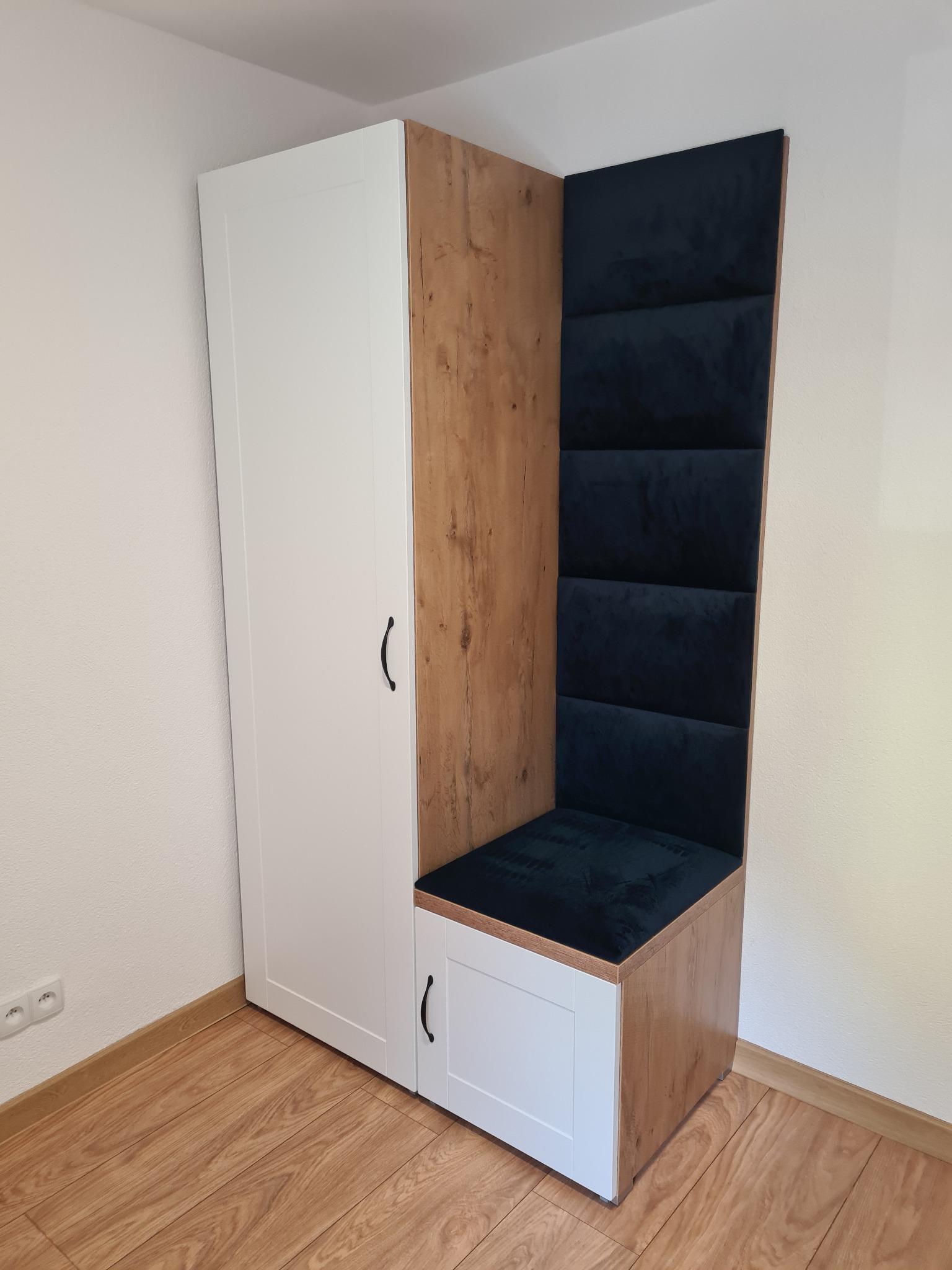 Biało-drewniana szafa z siedziskiem obitym granatowym welurem, wkomponowana we wnękę, z widocznymi gniazdkami elektrycznymi na ścianie i panelami podłogowymi.