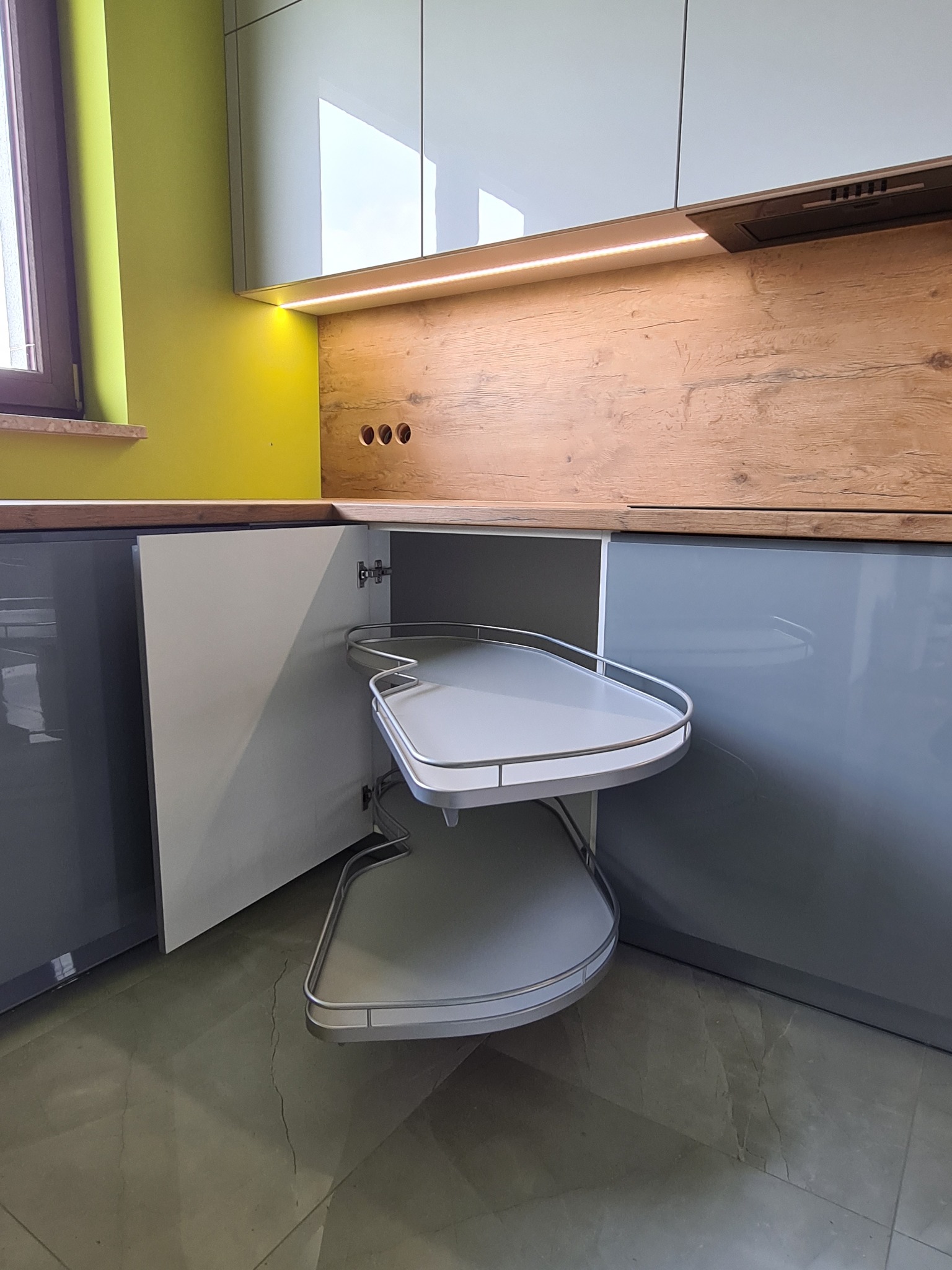 Wnętrze szafki narożnej z wysuwanymi półkami w nowoczesnej kuchni, widoczna drewniana ściana nad blatem i oświetlenie LED pod szafkami wiszącymi.