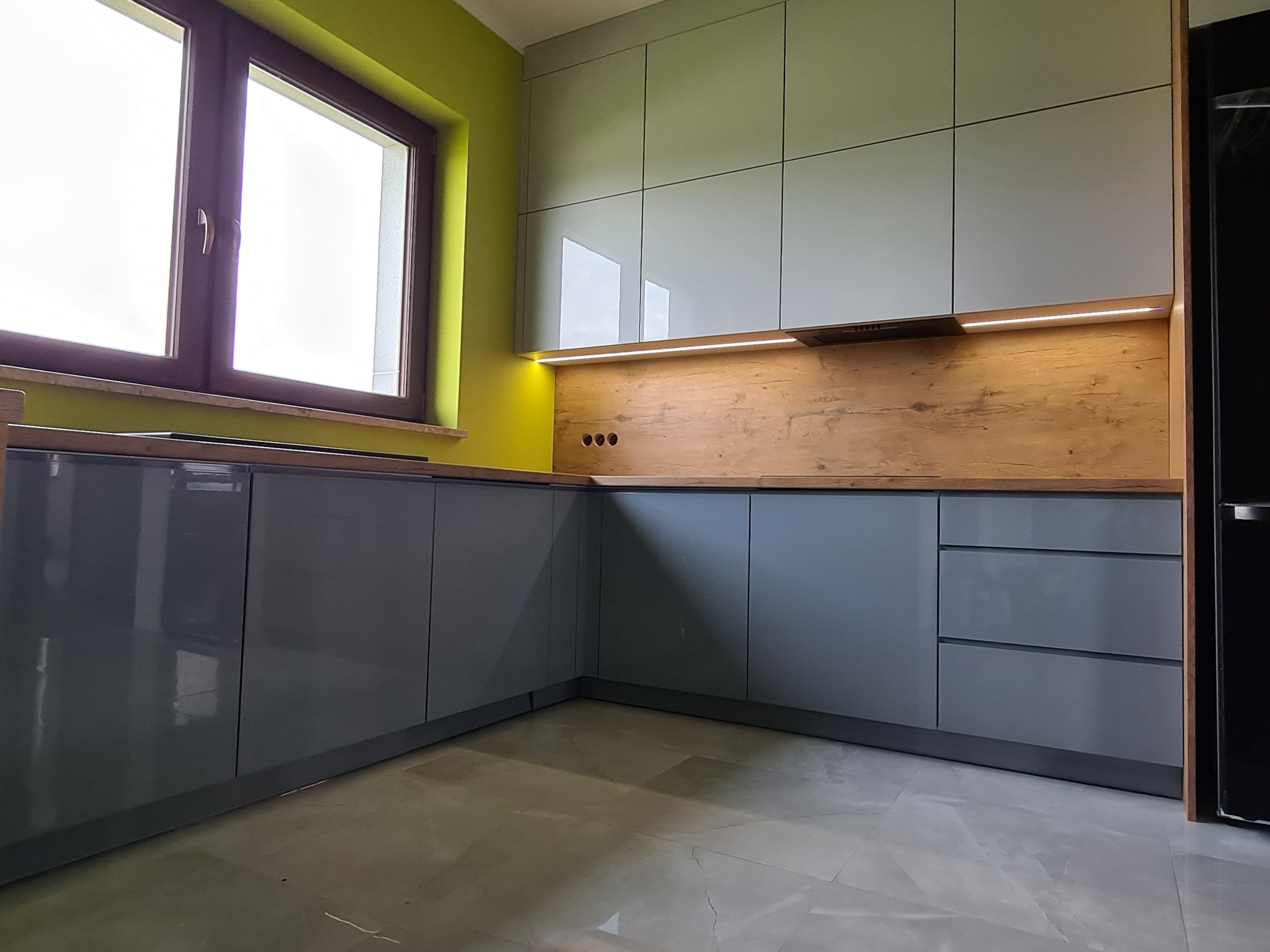 Nowoczesna zabudowa kuchenna w odcieniach szarości z drewnianym blatem i panelem ściennym, oświetlona listwą LED, na tle zielonej ściany i marmurowej podłogi.