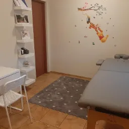 Gabinet logopedyczny z dziecięcym motywem Kubusia Puchatka na ścianie, białe biurko z krzesłem, półki i szary stół do masażu.