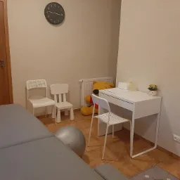 Gabinet w Kłodawie z szarym materacem rehabilitacyjnym, białym biurkiem, krzesłami dziecięcymi i pluszowym misiem Puchatkiem w tle.