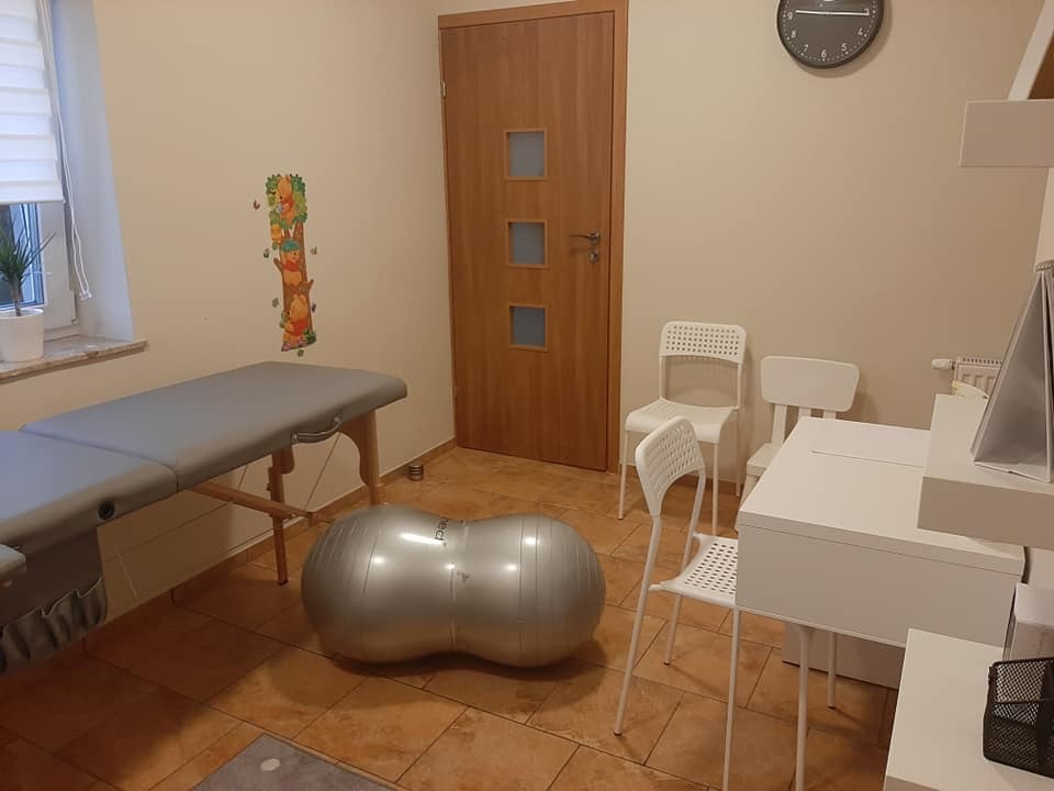 Gabinet terapeutyczny z leżanką, dużą szarą piłką rehabilitacyjną, białymi krzesłami i biurkiem, na ścianie naklejka z Kubusiem Puchatkiem.